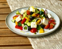 Fresh cucumbers, cherry tomatoes, bell peppers, red onions, black olives, capers, and creamy feta cheese, dressed with olive oil and oregano. Greek salad with cucumbers, tomatoes, olives, red onions, bell peppers, and feta cheese on a white plate